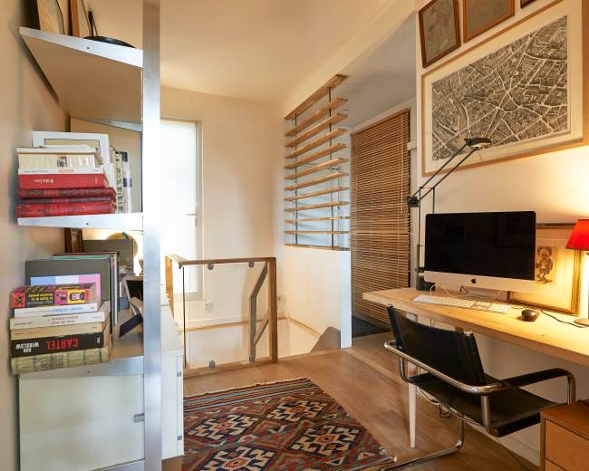 A modern home office features a wooden desk with an iMac, a stack of books on a shelf, and art framed on the walls.
