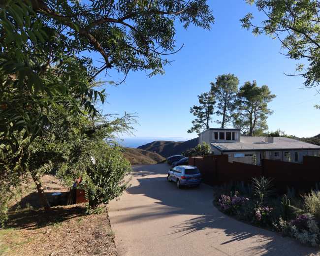 A house is situated on a hillside road overlooking the ocean, surrounded by trees and landscaped gardens.
