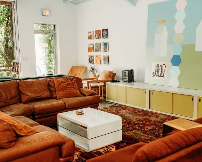 The image shows a cozy living area with brown couches, a coffee table, and an abstract mural on the wall, accompanied by decorative artwork and plants.