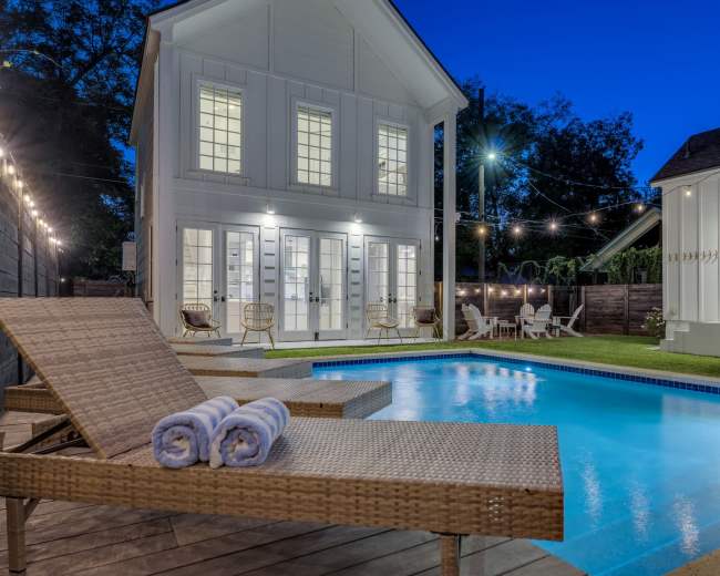 The image shows a modern house with a swimming pool and lounge chairs in a well-lit backyard during twilight.