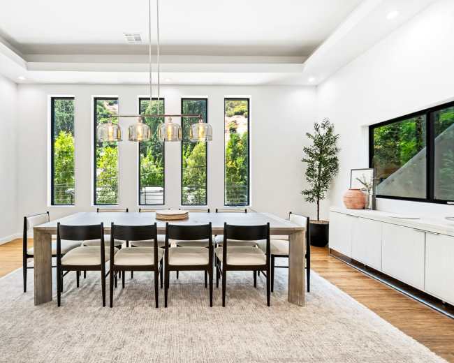 A modern dining room features a long table with eight chairs, surrounded by large windows that offer a view of greenery outside.