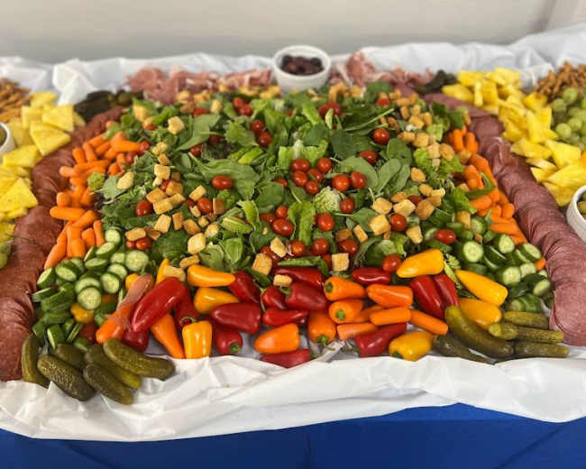 A large platter is arranged with an assortment of salad greens, cherry tomatoes, assorted peppers, pickles, olives, and various fruits, surrounded by bowls of grapes and crackers.