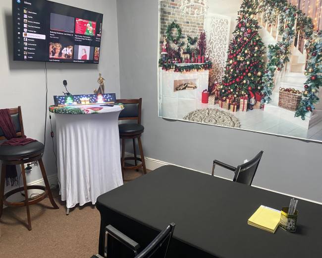The image shows a small, decorated room with a black table, chairs, a TV displaying a streaming interface, and a backdrop featuring a festive holiday scene.