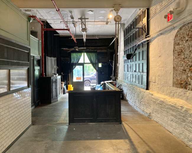 The image shows the interior of a vacant establishment with a bar area near the entrance, exposed brick walls, and green curtains visible through the front window.