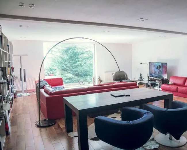 The image shows a modern living room with two red sofas, a large dining table, and a TV, all set against a backdrop of a large window with greenery visible outside.