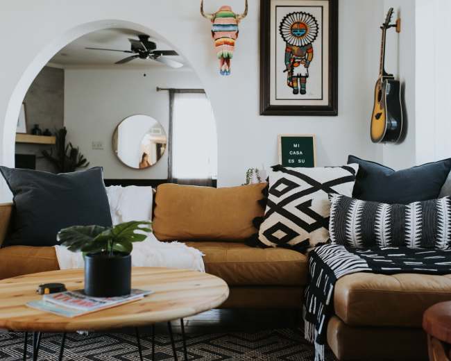 A cozy living room features a brown sofa with patterned pillows, a round wooden coffee table, and decor including artwork and a guitar.
