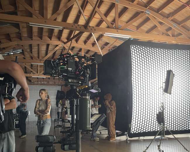 A film crew sets up equipment in a spacious studio, featuring cameras, lighting, and a figure in a costume behind a large softbox.