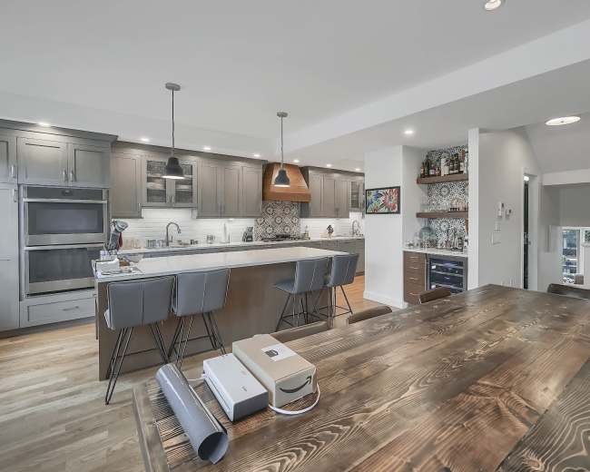 A modern kitchen features a large island with bar stools, wooden dining table, and stylish light fixtures.