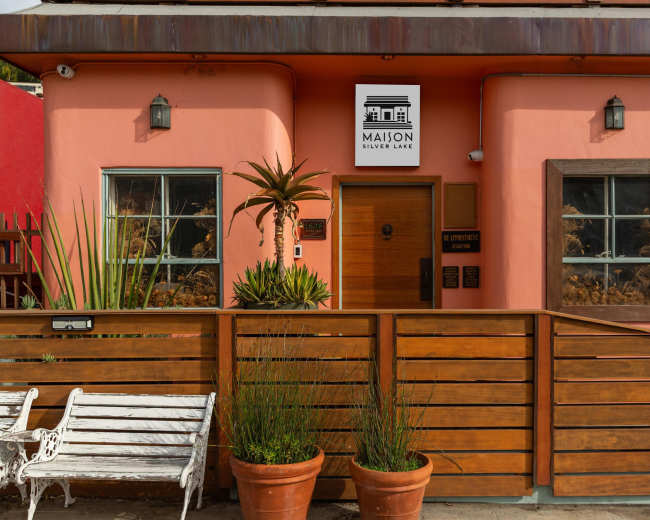 The image shows a pink building with a wooden entrance and a sign reading "MAISON SILVER LAKE," flanked by potted plants and a white bench in front.
