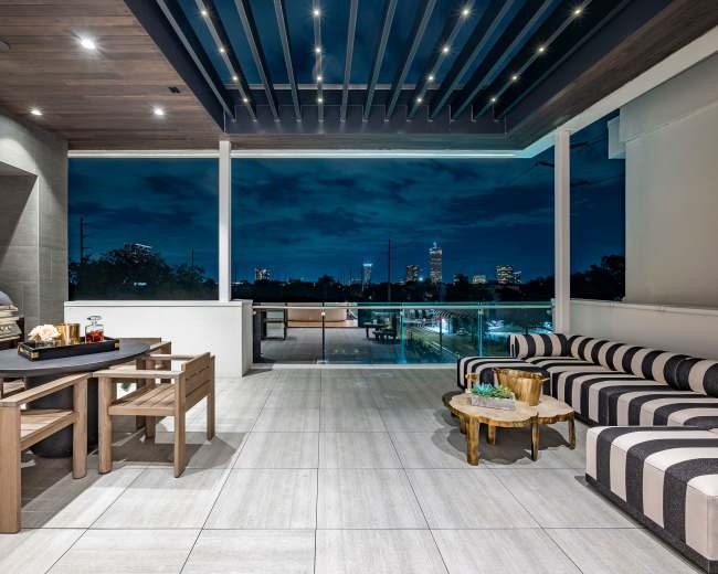 A modern outdoor patio with wooden seating, a dining set, a grill, and city skyline views at dusk.