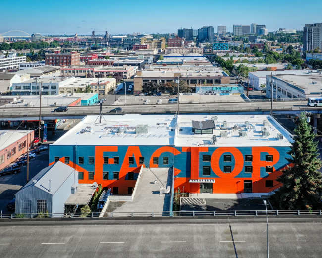 An industrial building with large, bold lettering painted on its exterior stands prominently against a city skyline featuring various commercial and residential structures.