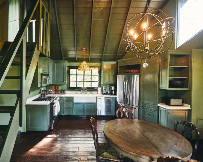 The image shows a rustic kitchen and dining area with green cabinetry, hardwood flooring, and a chandelier.