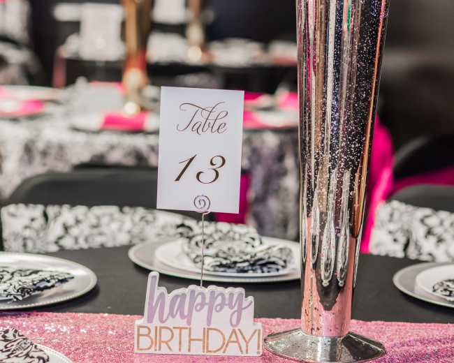 A decorated table setting features a centerpiece, a card displaying "Table 13," and a sign that reads "happy BIRTHDAY."