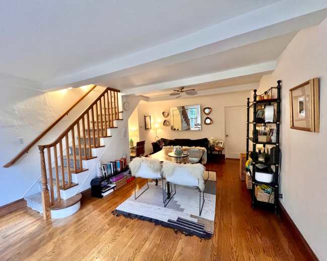 The image shows a spacious living area with a staircase, a seating arrangement, and a bookshelf against the wall.