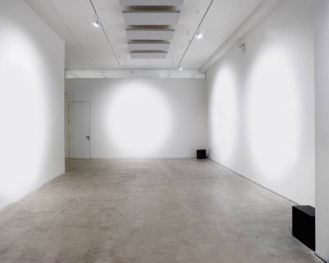 The image depicts a spacious, empty art gallery with white walls, a concrete floor, and minimal lighting fixtures.