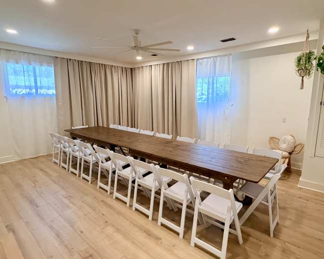 The image shows a spacious meeting room with a long wooden table surrounded by white folding chairs, large windows, and light-colored curtains.