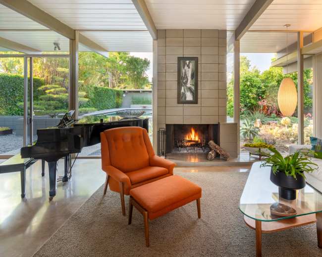 A modern living room features a fireplace, an orange armchair, a glass coffee table, and large windows overlooking a landscaped garden.
