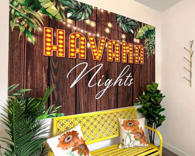 The image shows a brightly decorated corner featuring a yellow bench with floral cushions, a vibrant mural reading "HAVANA Nights" with lights, and plants in a well-lit interior space.