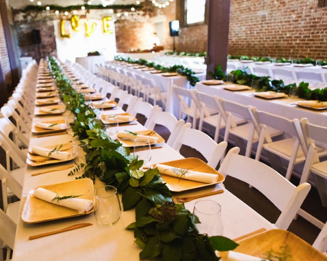 A row of elegantly set tables with white chairs is adorned with greenery and wooden serving trays, arranged for a dining event in a rustic venue.