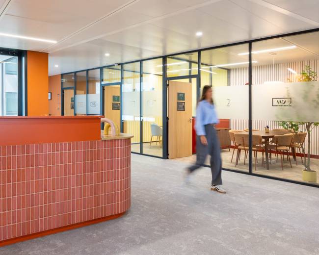 A person walks past a reception area in an office hallway featuring glass-walled meeting rooms and modern decor.