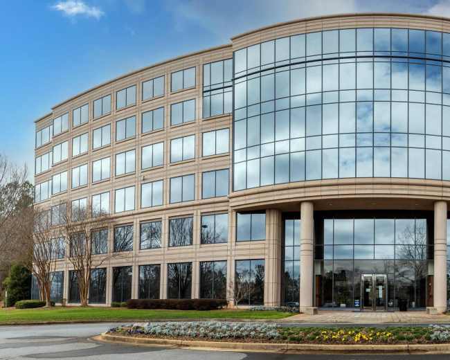 The image shows a modern office building with large glass windows and a curved façade, surrounded by landscaped greenery and flowers.