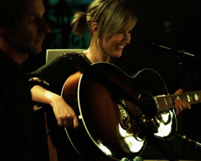 A woman plays an acoustic guitar while smiling, as a man stands nearby in a dimly lit setting.