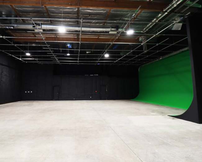 The image shows a spacious studio with a green screen on one side and black walls, featuring overhead lighting and a concrete floor.