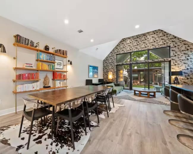 A spacious modern dining area features a long wooden table surrounded by black chairs, with bookshelves along one wall and large windows overlooking a landscaped outdoor space.