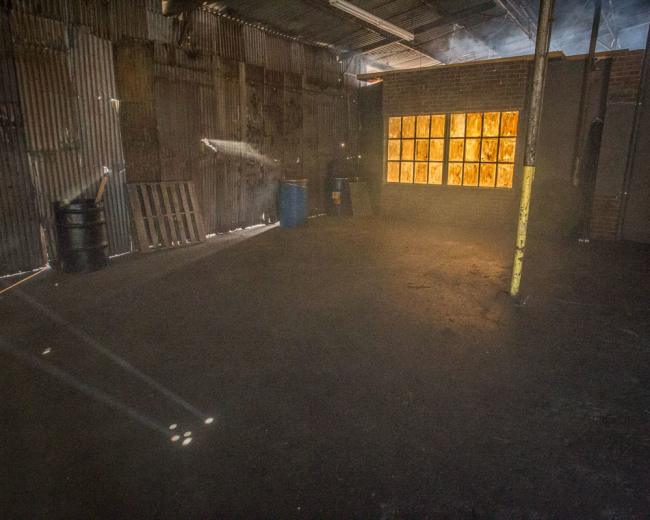 An empty industrial space features bare concrete floors, rusted metal walls, and a large window with wooden panes allowing light to stream in.