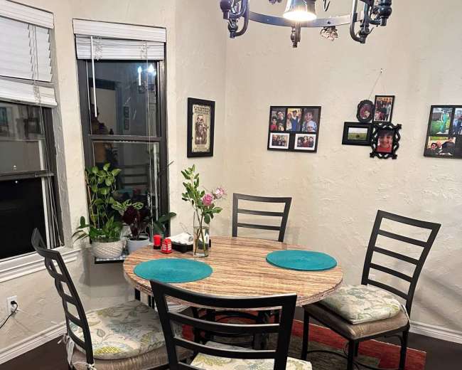 The image shows a round dining table surrounded by four chairs, with decorative placemats and a vase of flowers, all set against a backdrop of wall decor and a rug.