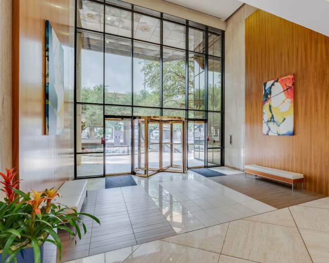 The image shows a modern lobby with large glass doors, a revolving door, wooden walls, and colorful artwork, flanked by potted plants.