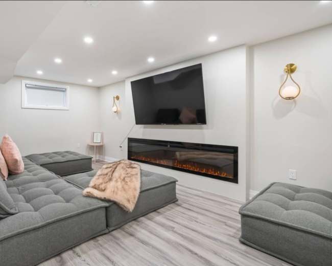 The image shows a modern living room with a gray sectional sofa, a wall-mounted TV, and an electric fireplace beneath.