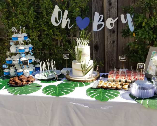A dessert table is set up outdoors, featuring blue cupcakes, a white cake, and a variety of treats arranged on a green tablecloth with tropical leaves, complemented by a decorative "Oh Boy!" banner.
