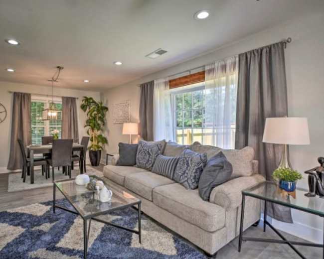 The living room features a light-colored sofa with decorative pillows, a glass coffee table, and a dining area with a chandelier visible in the background.