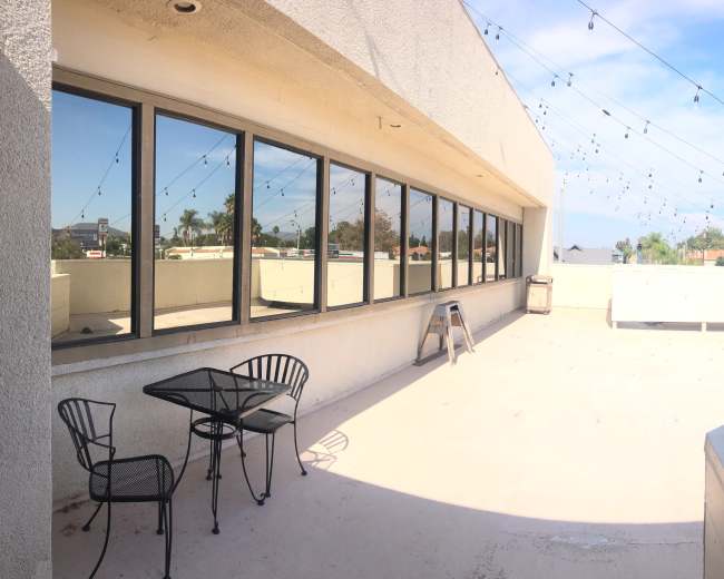 A panoramic view of a rooftop terrace featuring a table and chairs, with strings of lights overhead and a clear blue sky in the background.