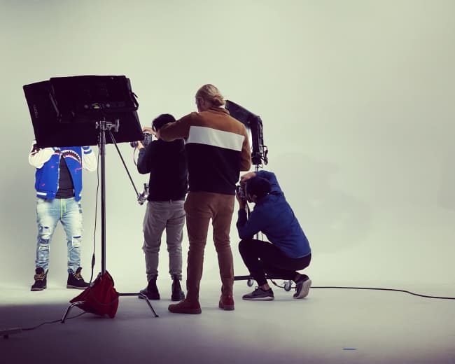 A group of three people is preparing a photoshoot in a studio setting with a plain white backdrop and studio lighting equipment.