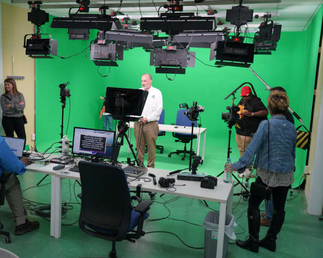 A video production studio with green screen setup, equipped with cameras, lighting, and crew members preparing for filming.