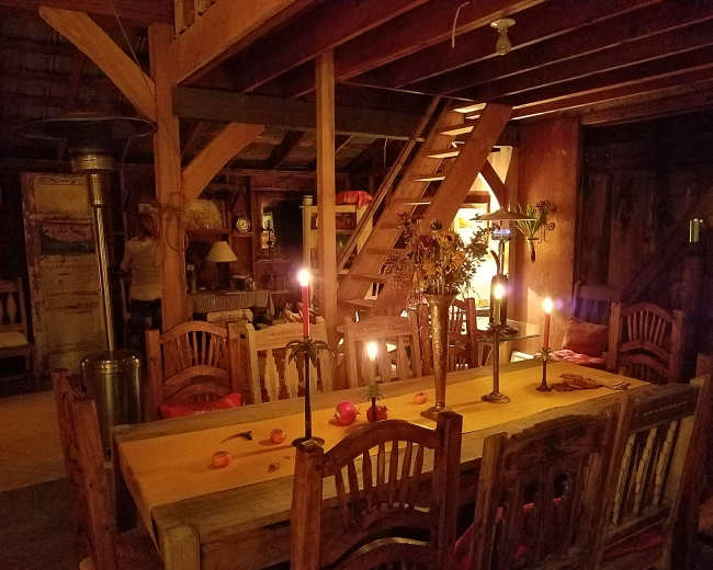 A warmly lit wooden dining area with a long table surrounded by chairs, a staircase in the background, and candles providing soft illumination.