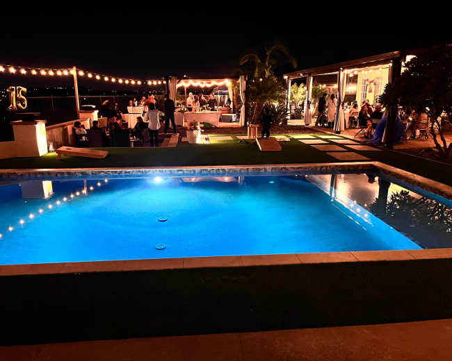 A brightly lit pool area is set for a nighttime celebration with string lights overhead and guests mingling by a decorated outdoor space.