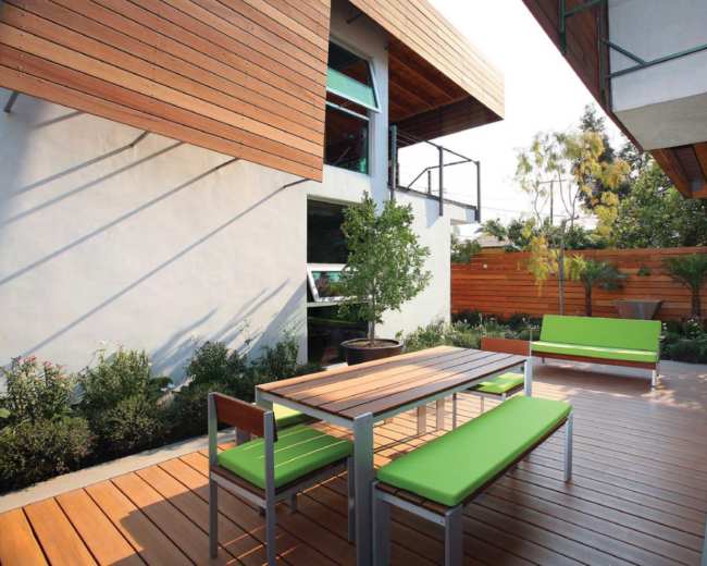 The image shows a modern outdoor patio with a wooden table and green benches surrounded by landscaped plants and wooden fencing.