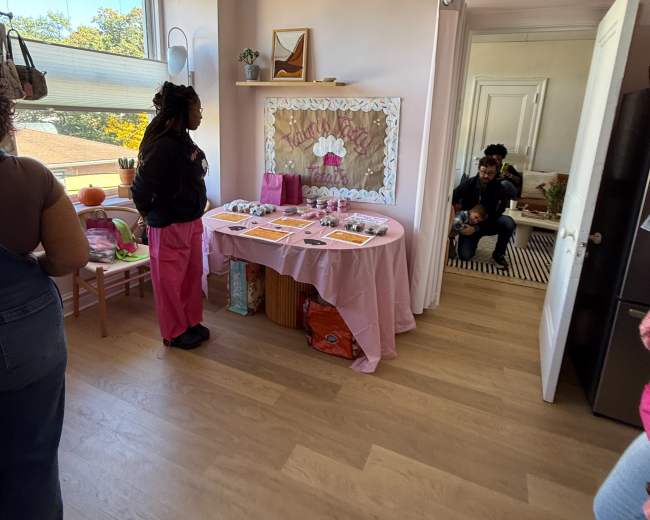 The image shows a decorated room with a table covered in pink cloth and various items displayed on it, while several people engage in conversation.