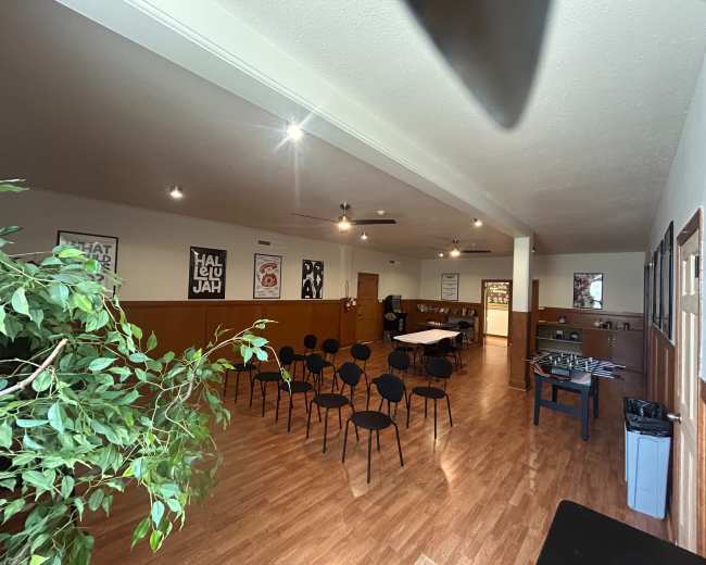 The image shows a spacious indoor room set up with black chairs facing a table, complemented by posters on the walls and a potted plant in the foreground.