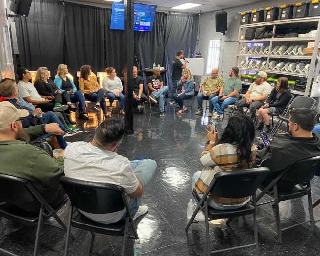 A group of people is seated in a circular arrangement in a well-lit meeting room with a presentation screen in the background.