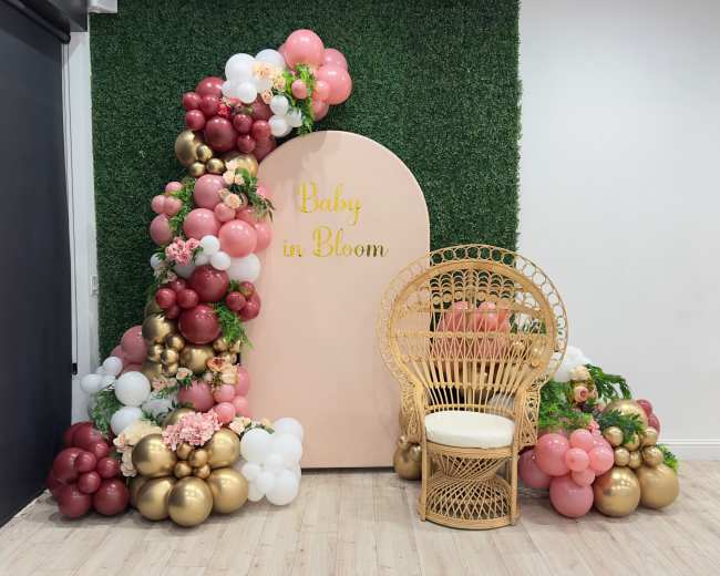 The image shows a decorative setup featuring a pink backdrop with the words "Baby in Bloom," adorned with colorful balloons and a rattan chair placed in front.