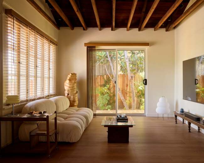 The image depicts a modern living room with a large window opening to a garden, featuring a plush sofa, a wooden coffee table, and a television mounted on the wall.