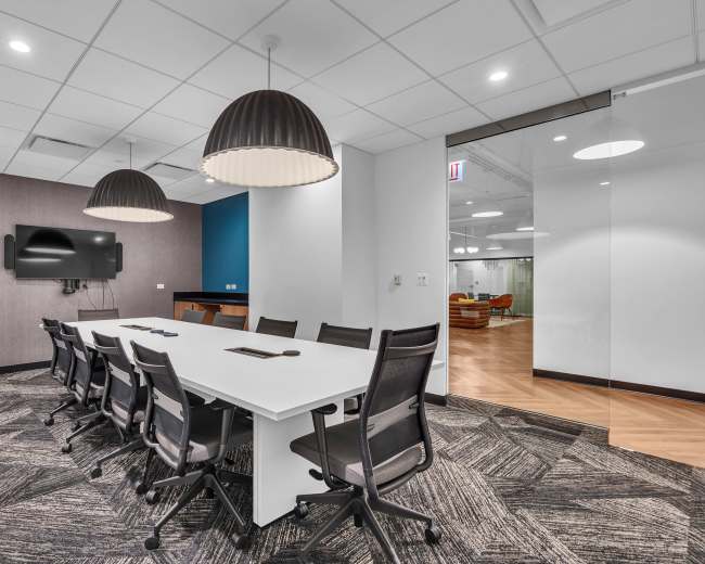 A modern conference room features a long white table surrounded by black chairs, with two large pendant lights above and a wall-mounted television.