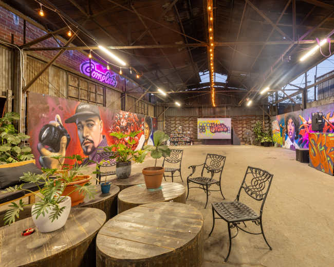 The image shows an industrial-style event space featuring graffiti murals on the walls, plants in pots, and wooden round tables surrounded by metal chairs.