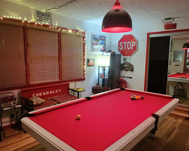 A game room featuring a red pool table, a stop sign, and various decorations, with warm lighting and a mirror reflecting part of the space.