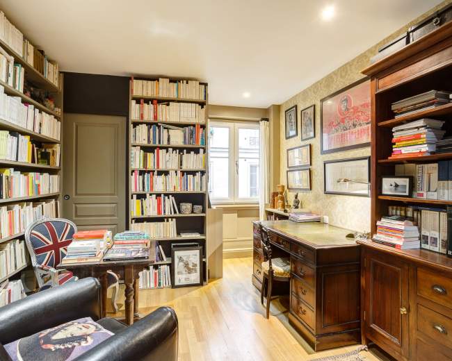The image shows a small, organized study with bookshelves filled with neatly arranged books, a wooden desk, and a chair featuring a Union Jack design.