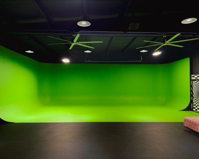 A studio with a bright green backdrop and a black floor, illuminated by ceiling lights and fans.